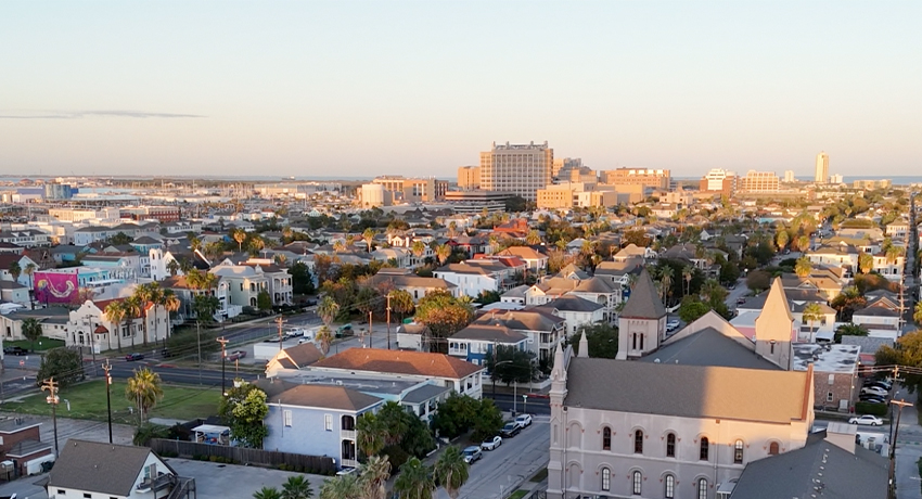 Aerial view of a coastal cityscape at sunset, showing residential blocks, tree‑lined streets, and larger buildings near the shoreline in the distance.