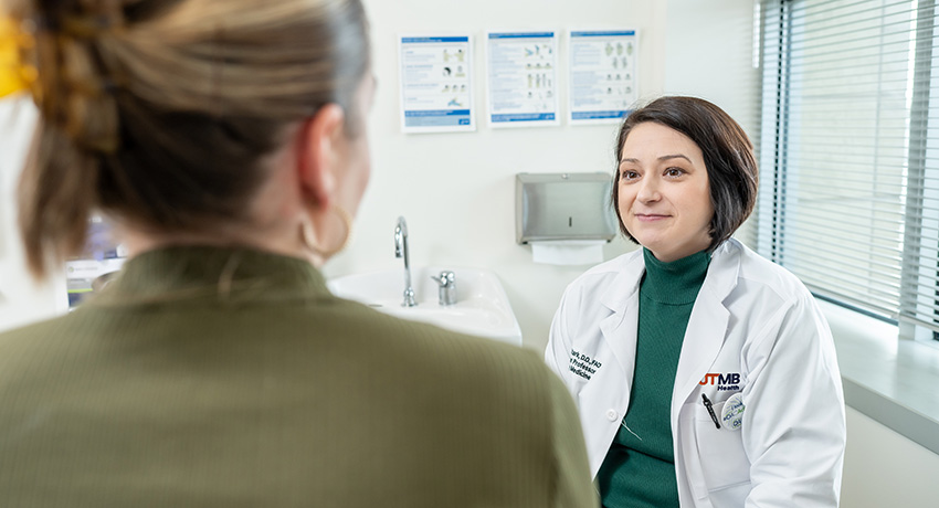 Health care provider in a white coat seated in an exam room, speaking with a patient whose back is to the camera. Medical posters, a sink, and a window with blinds are visible in the room.