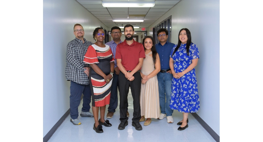 A group of seven healthcare professionals stands together in a hospital hallway, posing for a group photo. The group includes four men and three women, dressed in a mix of professional and casual attire.