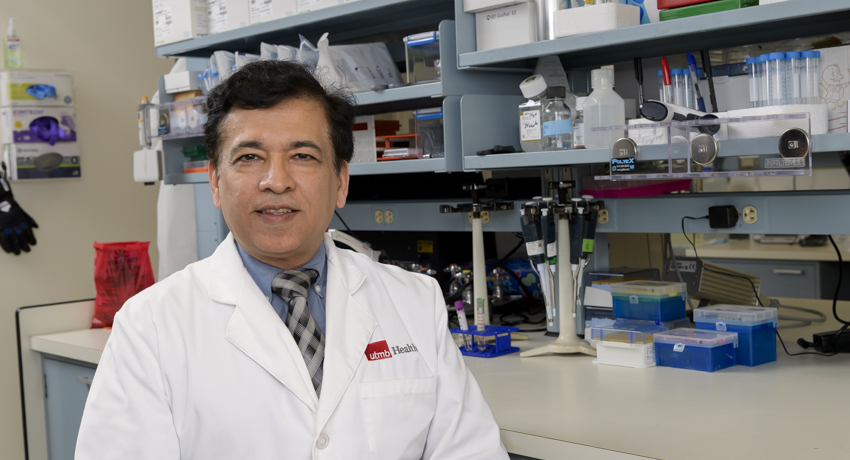 A man wearing a white UTMB Health lab coat sits in a biomedical research laboratory, facing the camera. Shelves behind him hold pipettes, test tubes, laboratory supplies, and scientific equipment, highlighting the research environment.