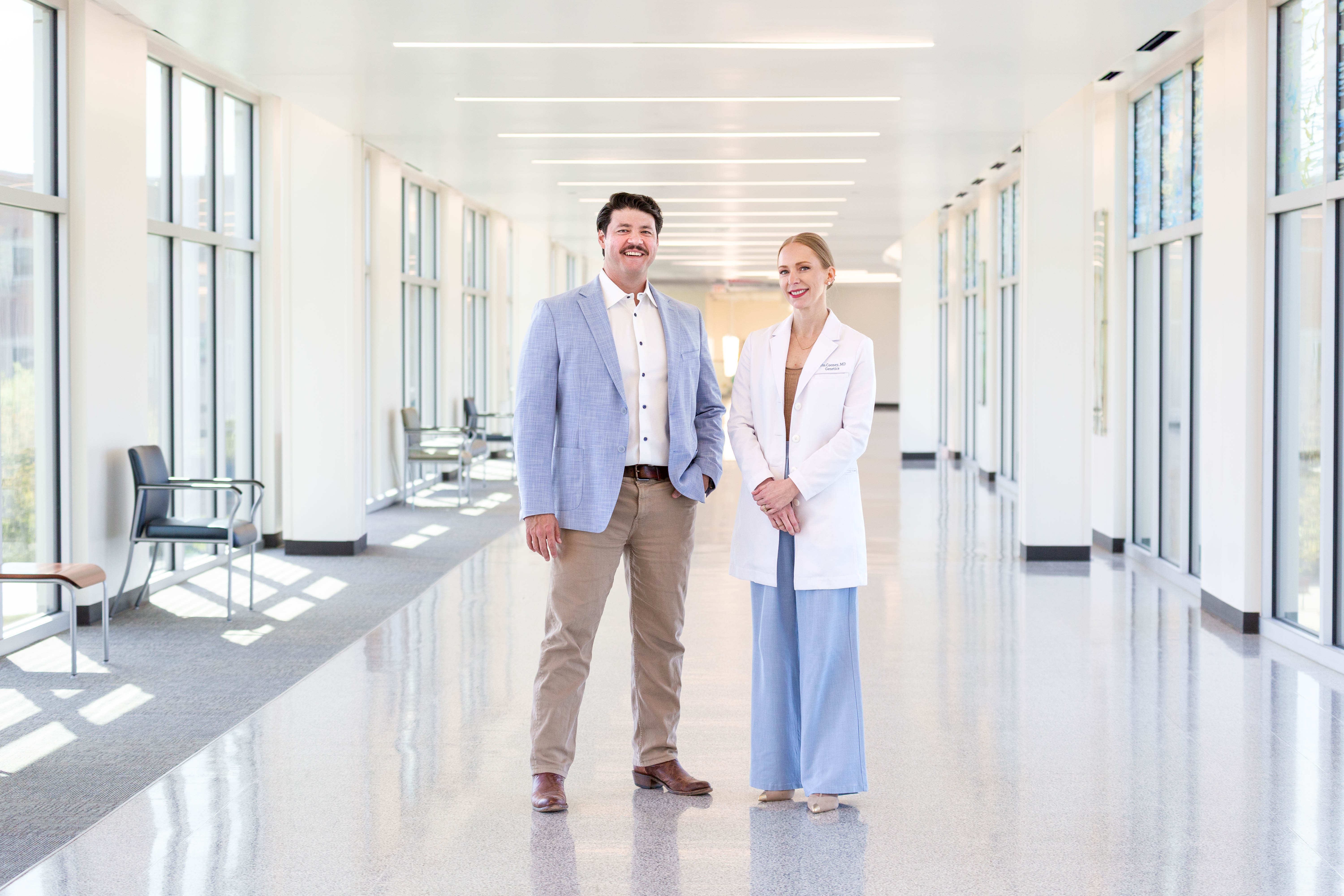 Two adults stand side by side in a bright hallway with large windows and overhead lighting, one wearing a white coat and the other a light-colored jacket.