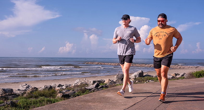 Two people jog on a paved path along a shoreline, with rocks, beach, ocean waves, and clouds visible in the background.