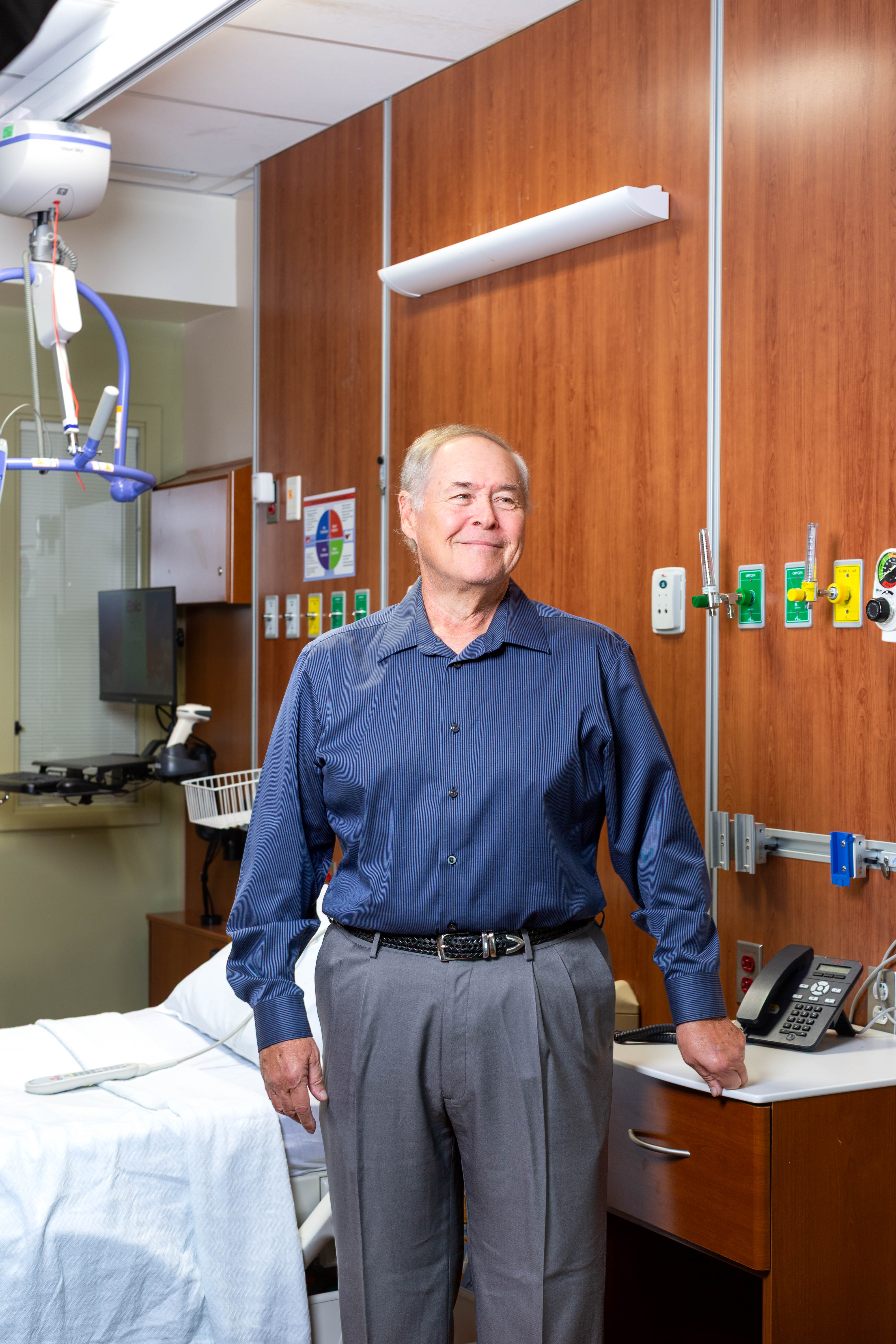 Older adult stands beside a hospital bed in a patient room, with wall‑mounted medical gas outlets, a ceiling lift, and a telephone visible nearby.
