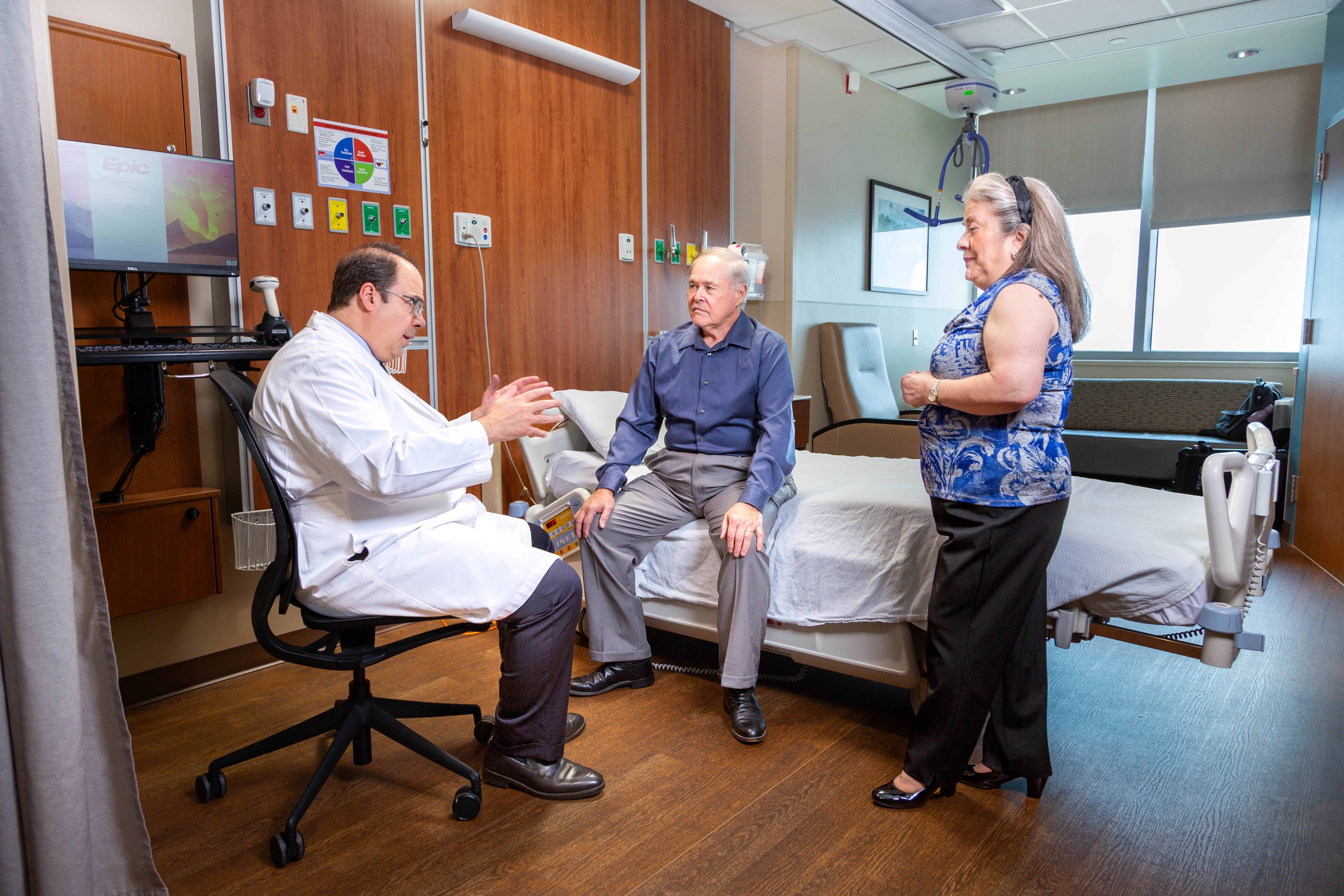 Clinician seated beside a patient on an exam bed speaks with the patient while another adult stands nearby in a clinical exam room with a computer workstation and wall‑mounted equipment visible.
