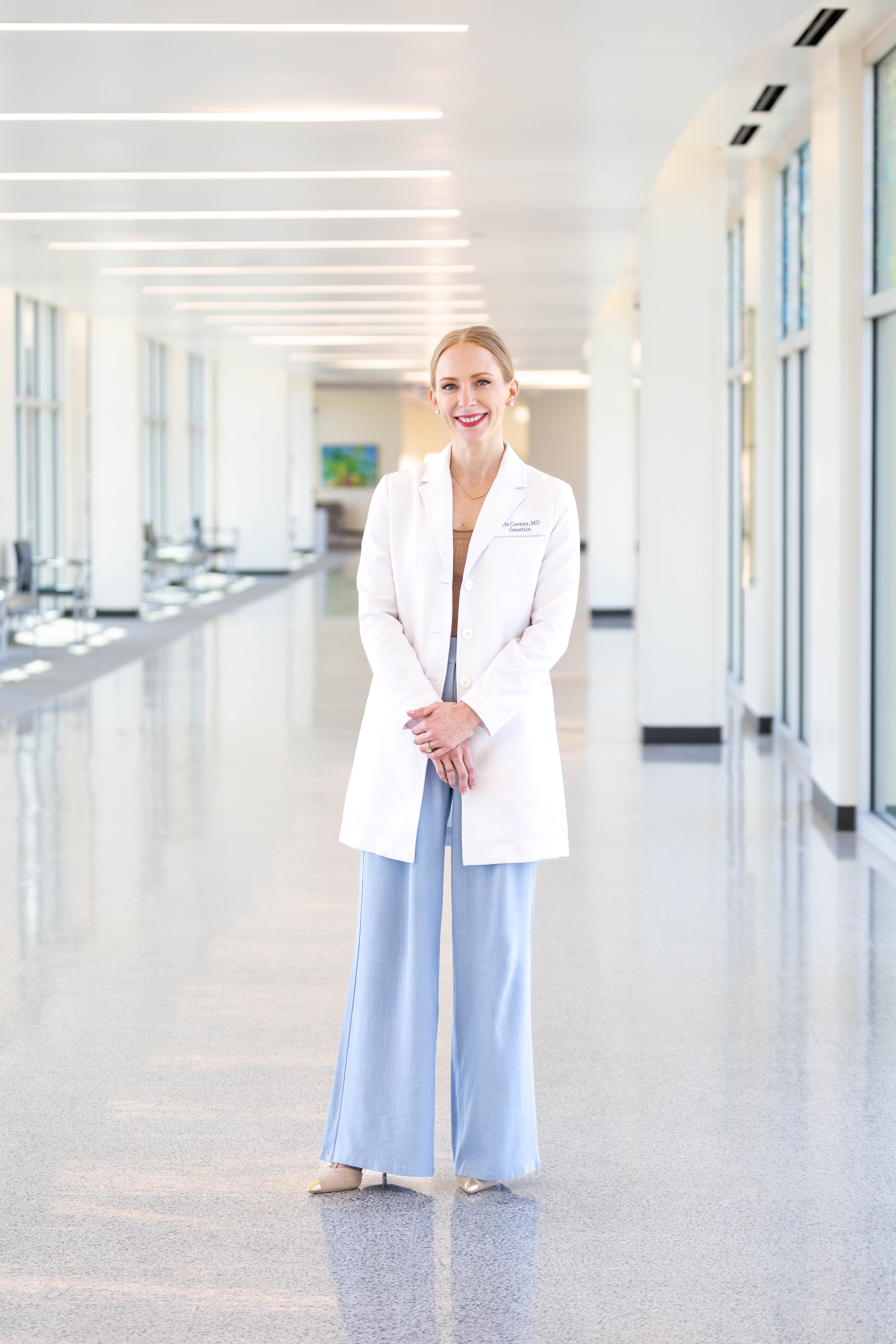 Clinician wearing a white coat stands in a bright hallway with large windows and overhead lighting, hands clasped in front.