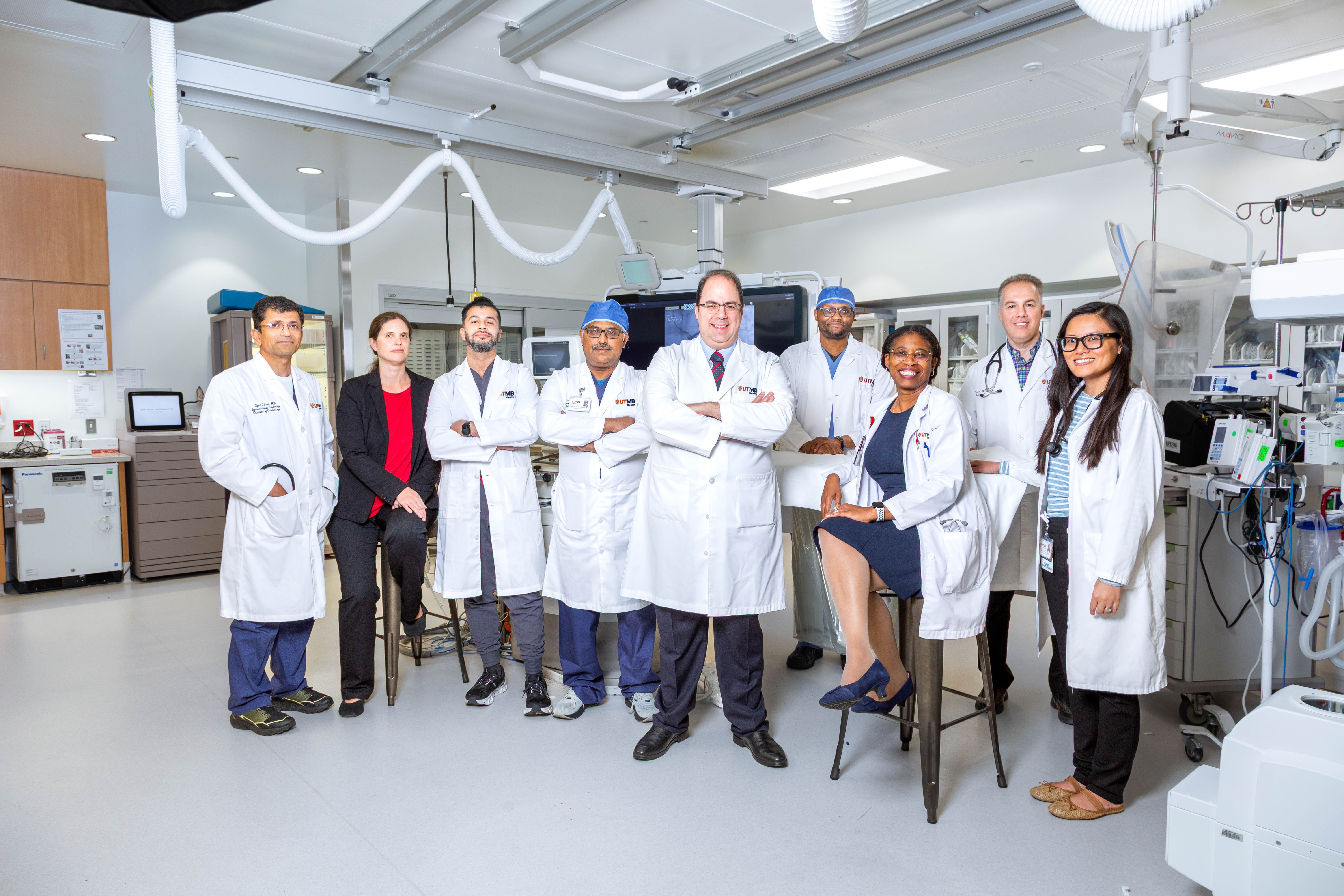 Group of clinicians in white coats stand together in a clinical procedure room with overhead equipment, monitors, and medical devices visible.