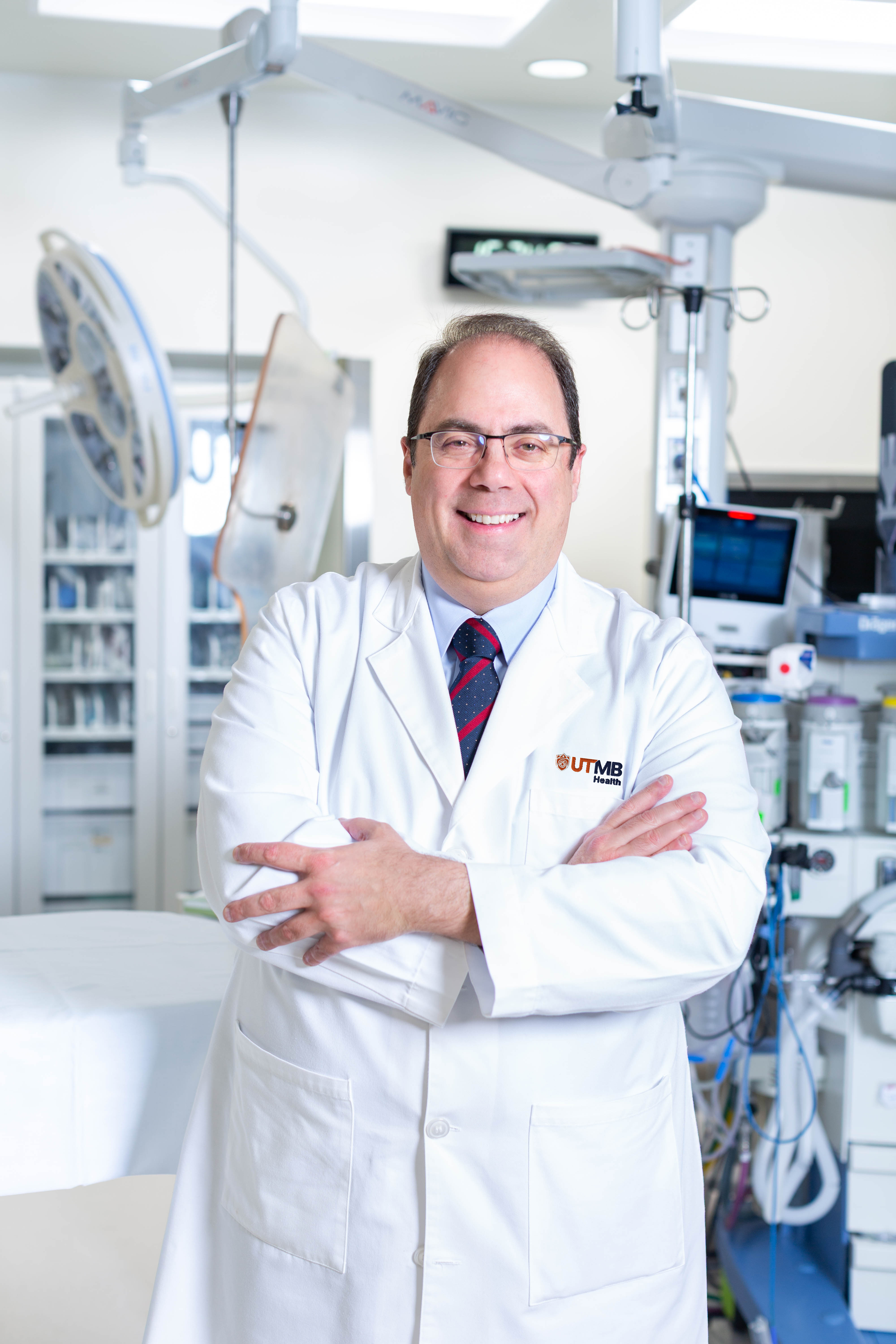 Clinician in a white coat with a UTMB Health logo stands with arms crossed in a clinical setting, with overhead equipment and monitors visible in the background.