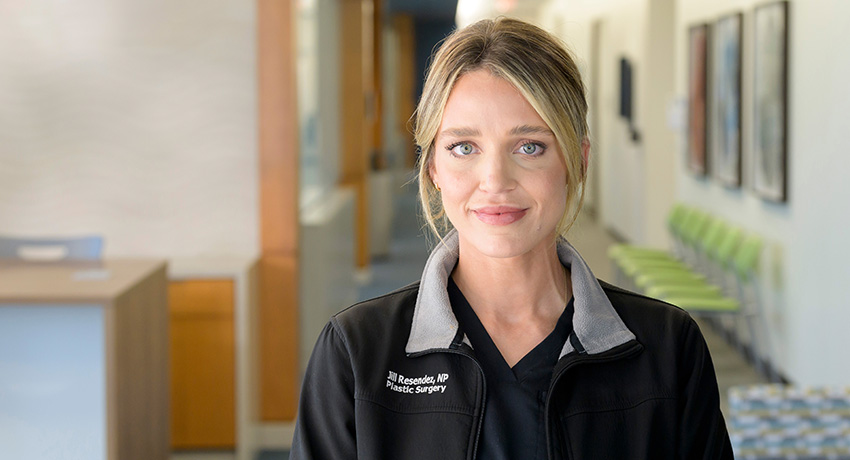 Portrait of a person wearing black medical scrubs and a black jacket, standing in a clinic hallway with seating visible in the background. The jacket is embroidered with “Jill Resendez, NP” and “Plastic Surgery.”