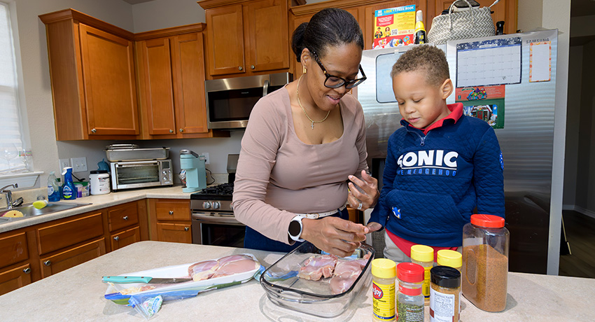 A preschooler standing at the kitchen island with his mother watches her season the chicken.