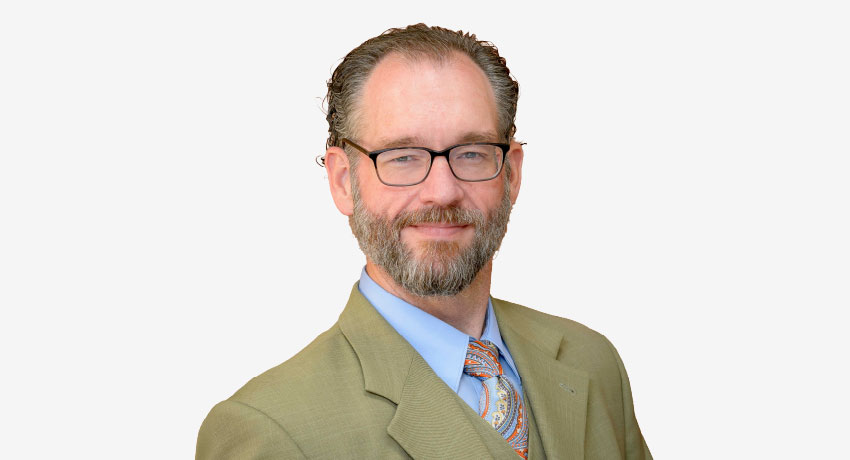 Head-and-shoulders portrait of a person wearing glasses, a light-colored suit jacket, blue shirt, and patterned tie against a plain light gray background. The subject is positioned in the center of the frame.
