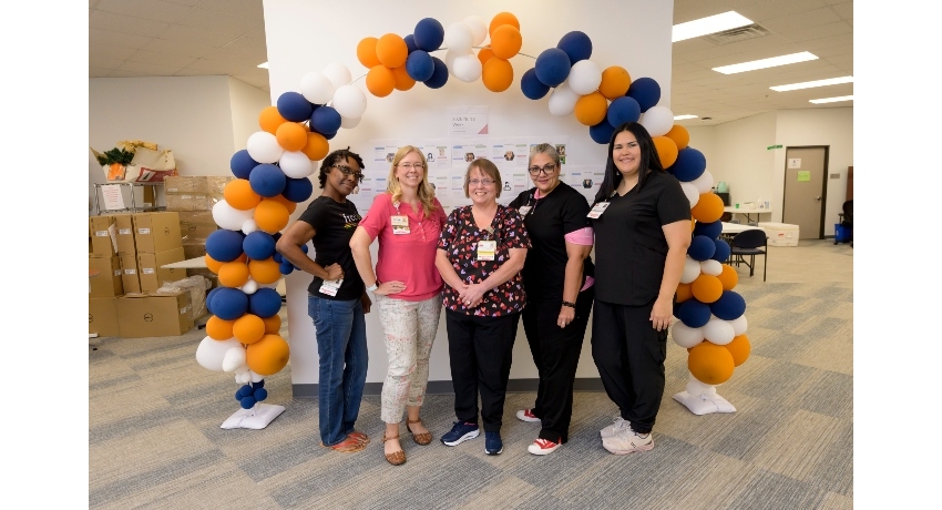 Five women wearing name badges stand together smiling beneath a balloon arch in blue, orange, and white inside a large office or clinical space. Boxes and supplies are visible in the background, suggesting an organized workspace or event setup.