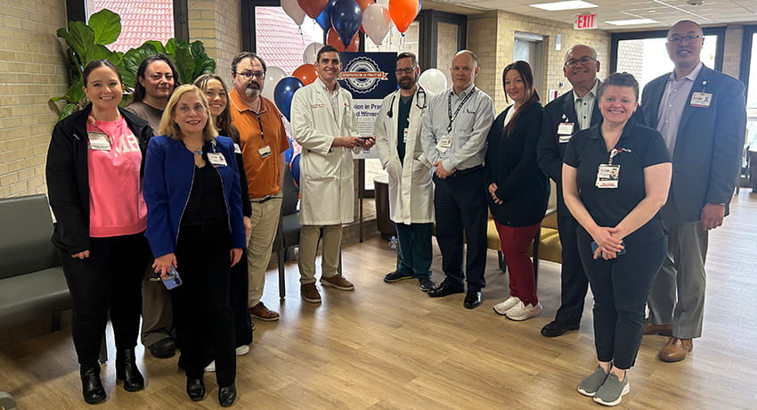Group of UTMB staff gathered indoors during an award presentation, standing near balloons and a sign recognizing team achievement.