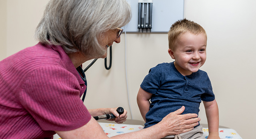 Healthcare provider examines a young child seated on an exam table in a medical exam room. Medical equipment is visible on the wall in the background.