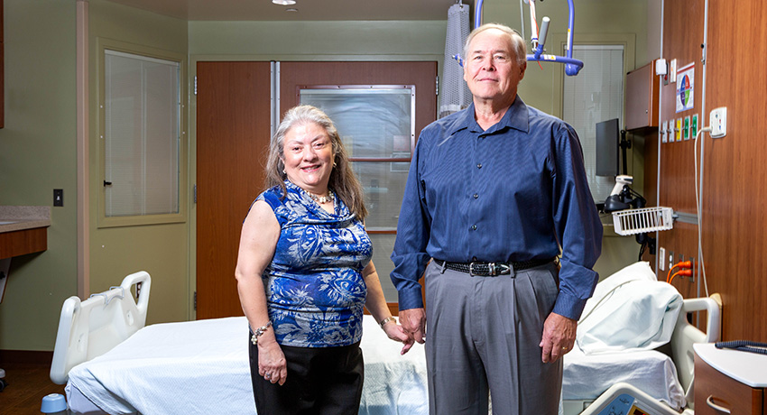Two adults stand side by side in a hospital room near a patient bed, with medical equipment and wall-mounted controls visible in the background.