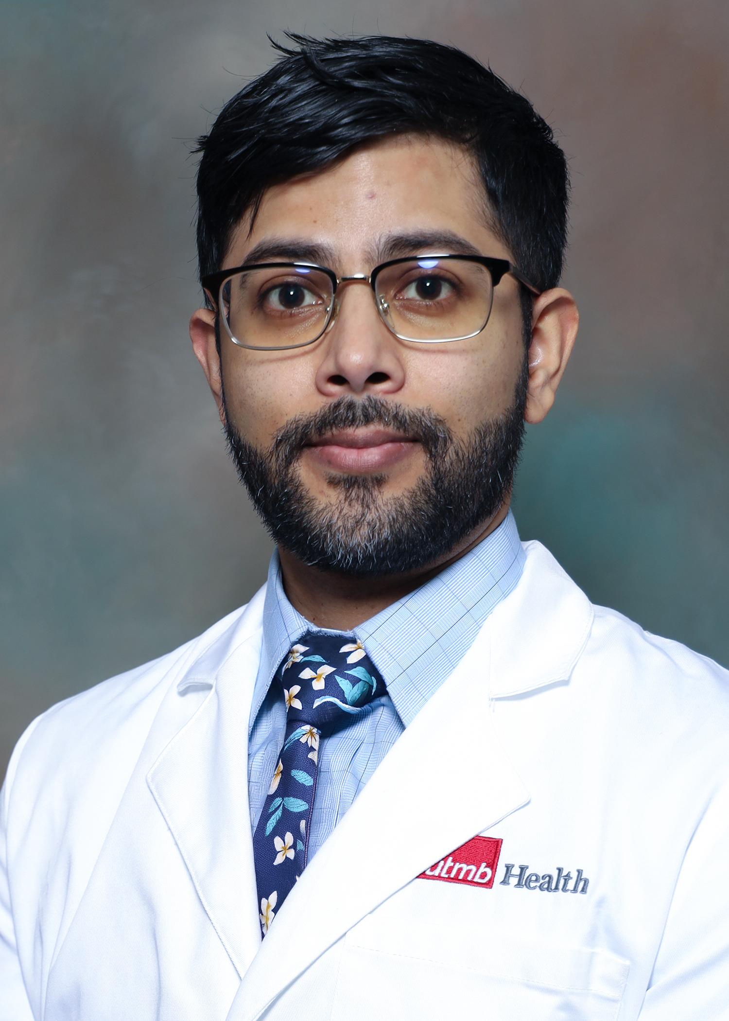 Person wearing a white lab coat with a UTMB Health logo, light blue dress shirt, and patterned tie against a neutral studio background.