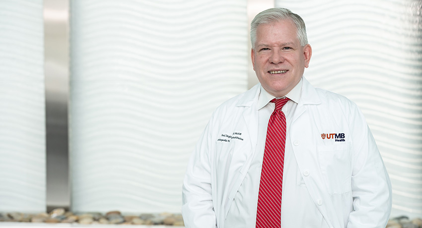 Person wearing a white UTMB Health lab coat and red striped tie stands in front of a light-colored textured wall. The UTMB Health logo is visible on the coat.