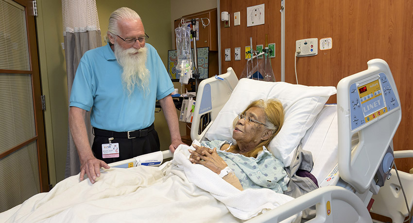 A volunteer stands next to a patient who is reclining in a hospital bed as the two of them chat.