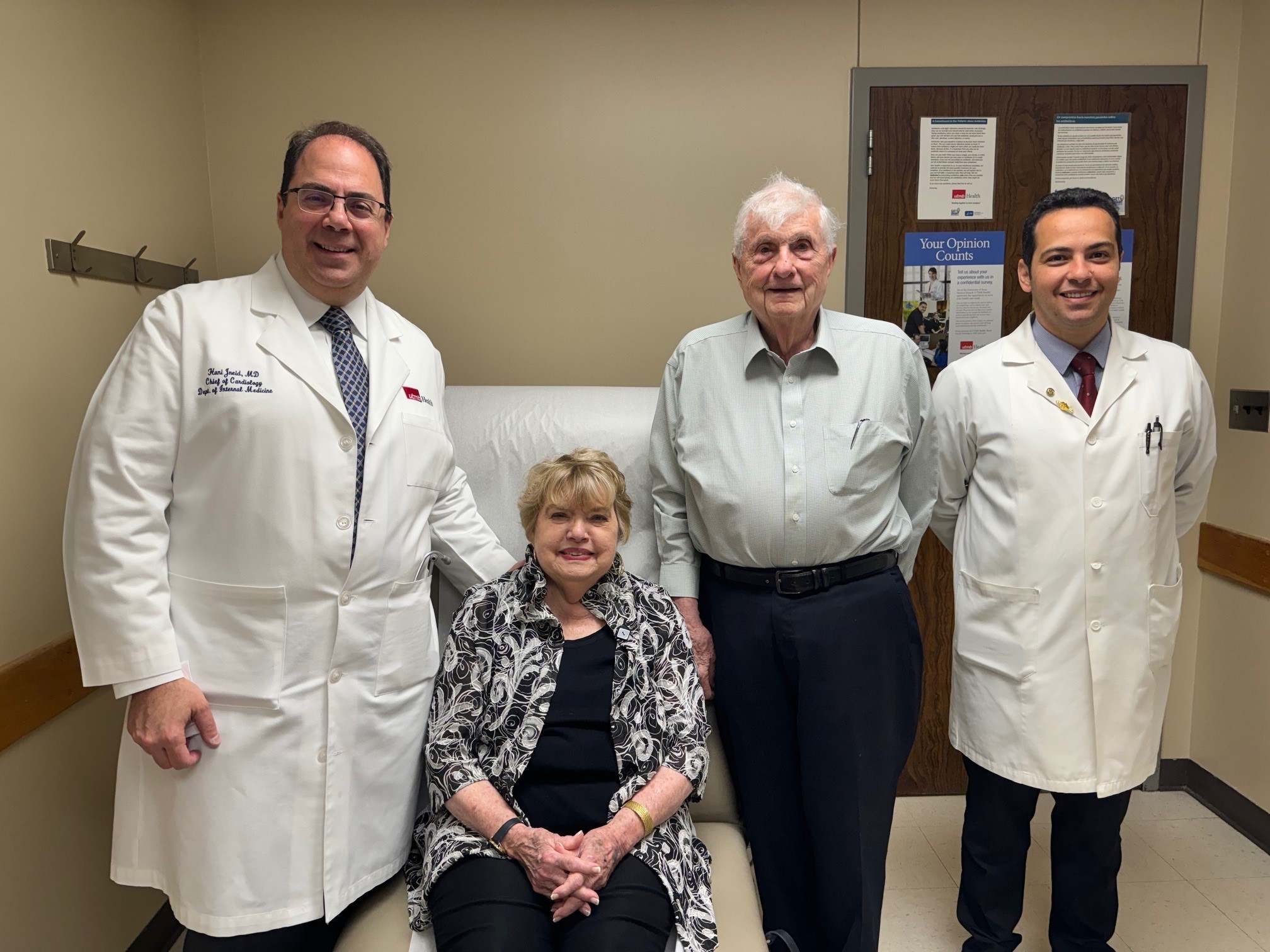 Four people standing and sitting in a medical exam room, including a physician in a white coat on the left and two others standing beside a seated patient.