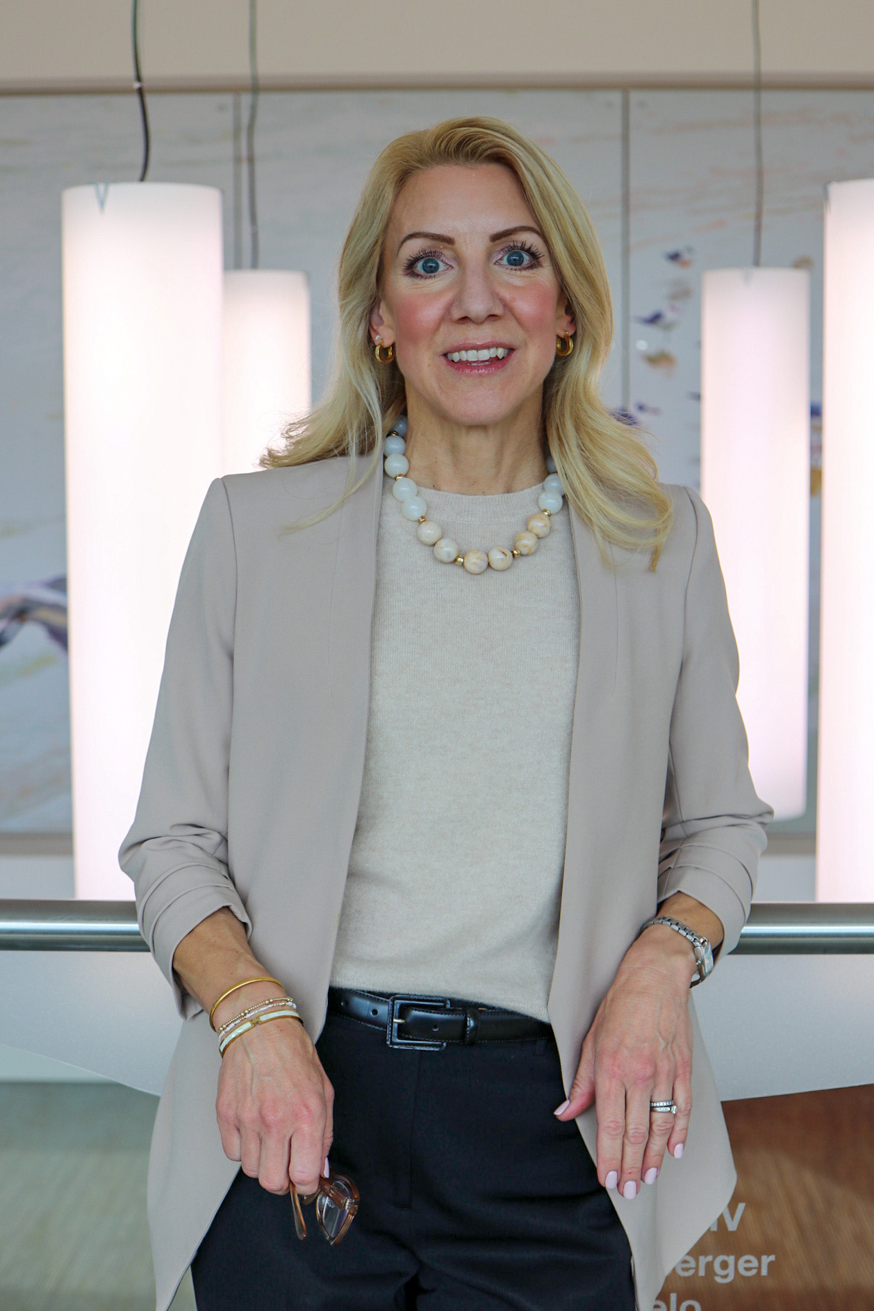 An individual standing indoors near a railing, dressed in a light blazer and dark pants, with illuminated vertical light panels and wall art in the background.