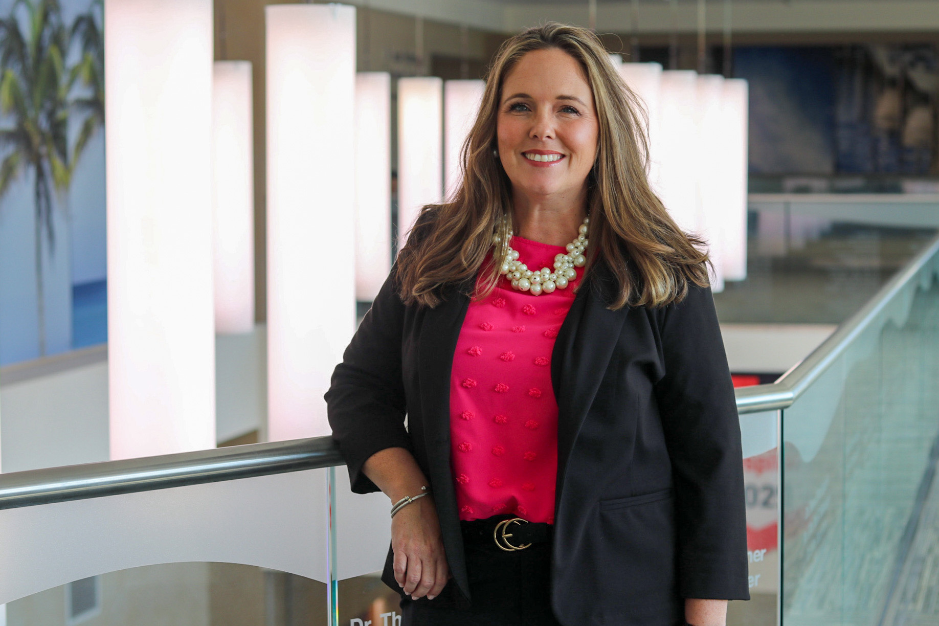 An individual standing indoors near a glass railing, wearing a dark jacket over a bright top with a large beaded necklace, with tall illuminated light panels in the background.