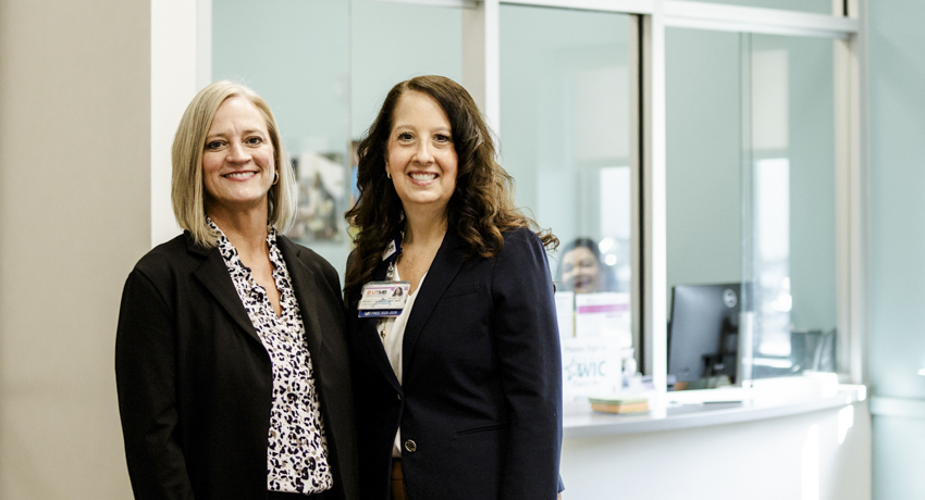 Tammy Jimenez and Aimee Jackson stand together at a UTMB Health WIC clinic, smiling for the camera near the clinic’s reception area.