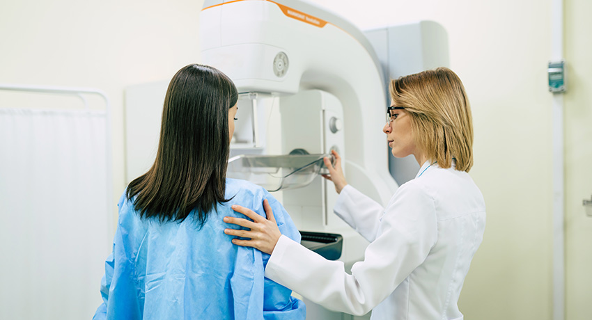 Patient in a blue medical gown stands at a mammography machine while a clinician in a white coat adjusts the equipment and positions the patient.