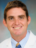  Man in this professional head-and-shoulder image has short brown hair and is wearing a white lab coat over a light blue shirt with a tie. 