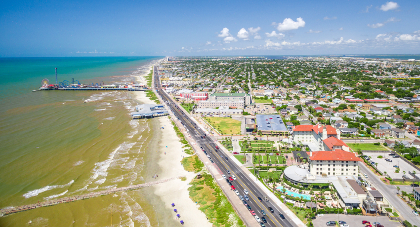 An aerial view of Galveston’s beachfront shows a shoreline road, sandy beach, Gulf waters, and a pier extending into the water, with a dense residential and commercial area stretching inland.