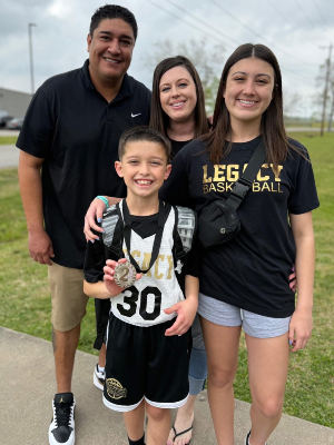 Jonah Martinez and his family, dad Jonathan, mom Jennifer and sister Jenna. Four people stand together outdoors on a sidewalk; a child in a sports jersey and backpack holds a medal in the foreground, with three other individuals behind on a grassy area.