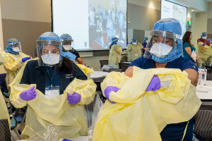 Healthcare workers wearing face shields, masks, and gloves secure yellow protective gowns in a classroom setting, with a presentation projected on a screen in the background.