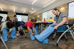 Participants in a classroom put on blue protective garments, with training materials and screens visible in the background.