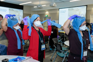 Healthcare staff in a training room adjust blue protective hoods over masks while seated at tables with supplies. Presentation slides and ceiling-mounted projectors are visible in the background.