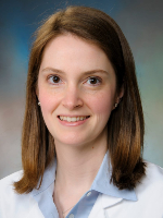  Professional headshot shows a woman wearing a white coat and light blue collared shirt against a blue-gray studio background..
