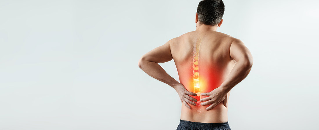 Person seen from behind stands against a light background, holding the lower back with both hands. A glowing red overlay highlights the spine and lumbar region, illustrating back pain or spinal stenosis.