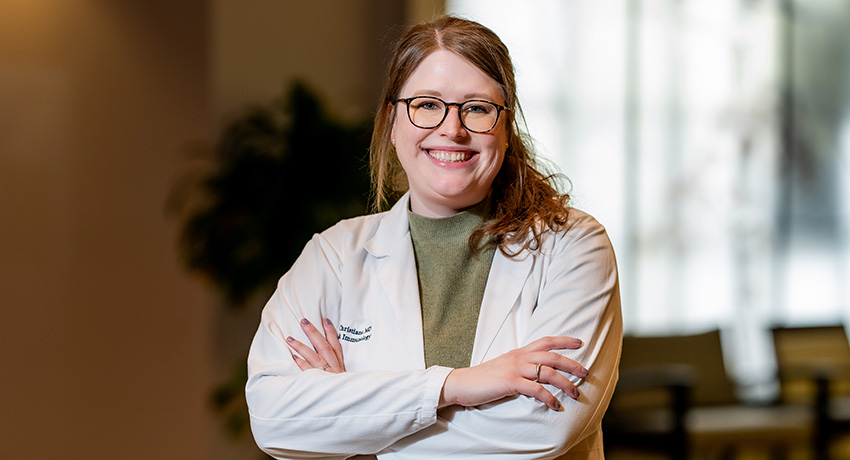 A woman wearing a white lab coat and glasses is standing indoors, with a blurred background that includes a plant and soft lighting.
