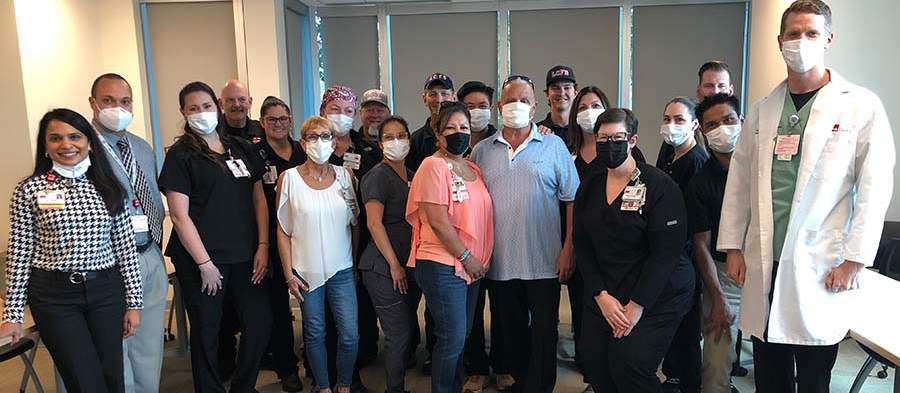 A group of UTMB Health emergency and urgent care doctors, nurses and staff stand together indoors with a heart attack survivor, smiling and wearing hospital badges and masks, recognizing the team that provided life‑saving care