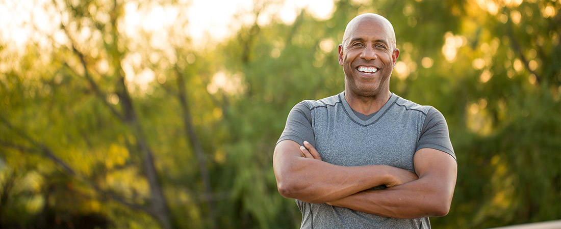 Person standing outdoors with arms crossed, wearing a short‑sleeve gray shirt, with trees and sunlight in the background